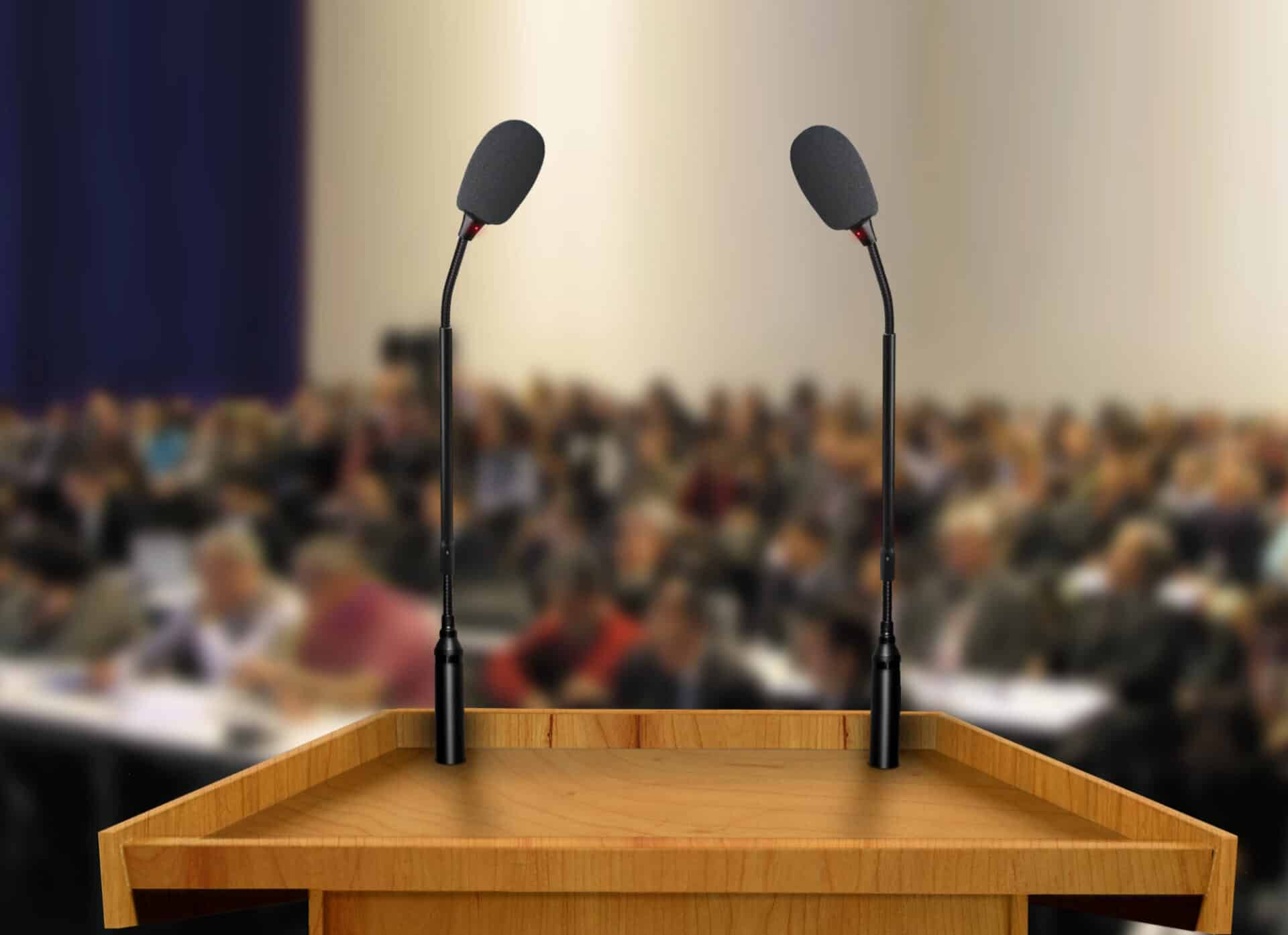 Podium with microphones in focus, blurred crowd of people in background, representing political marketing events and campaign strategies.